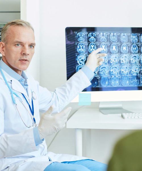 Portrait of handsome mature doctor pointing at brain scan images while consulting young woman in modern neurology clinic, copy space