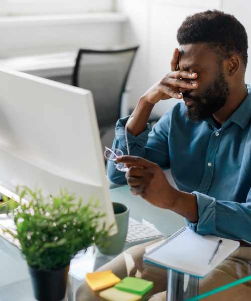 Tired black businessman feeling pain eyestrain, holding glasses and rubbing dry irritated eyes fatigued from computer work, man suffering from sight problem, sitting at workplace